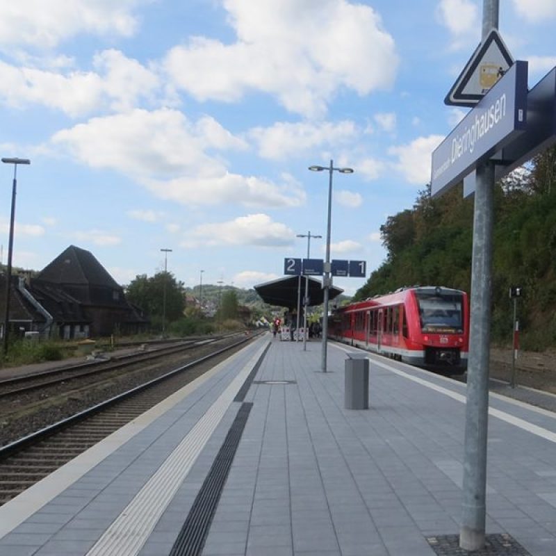 Bahnhof mit Gleisen und einem roten Zug, der am Bahnsteig 2 hält, umgeben von einer ländlichen Umgebung und leicht bewölktem Himmel
