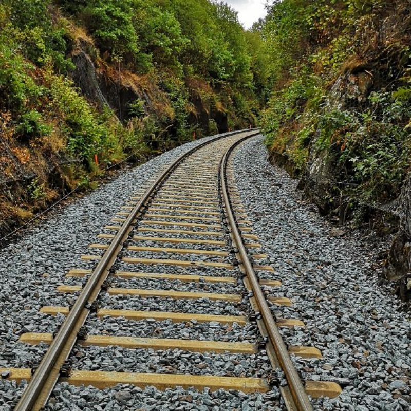 Eisenbahnschienen winden sich durch eine grüne Landschaft, umgeben von Felsen und Bäumen, auf einem mit Kies bedeckten Gleisbett