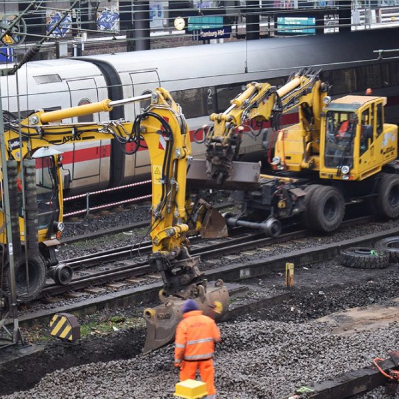 Zwei gelbe Baugeräte arbeiten an Bahngleisen, während ein Zug im Hintergrund vorbeifährt und ein Arbeiter in orangefarbener Kleidung steht