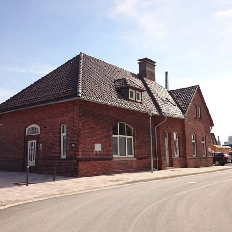 Ein rotes Backsteingebäude mit einem Satteldach, umgeben von einer Straße und mehreren Fahrzeugen, bei sonnigem Himmel