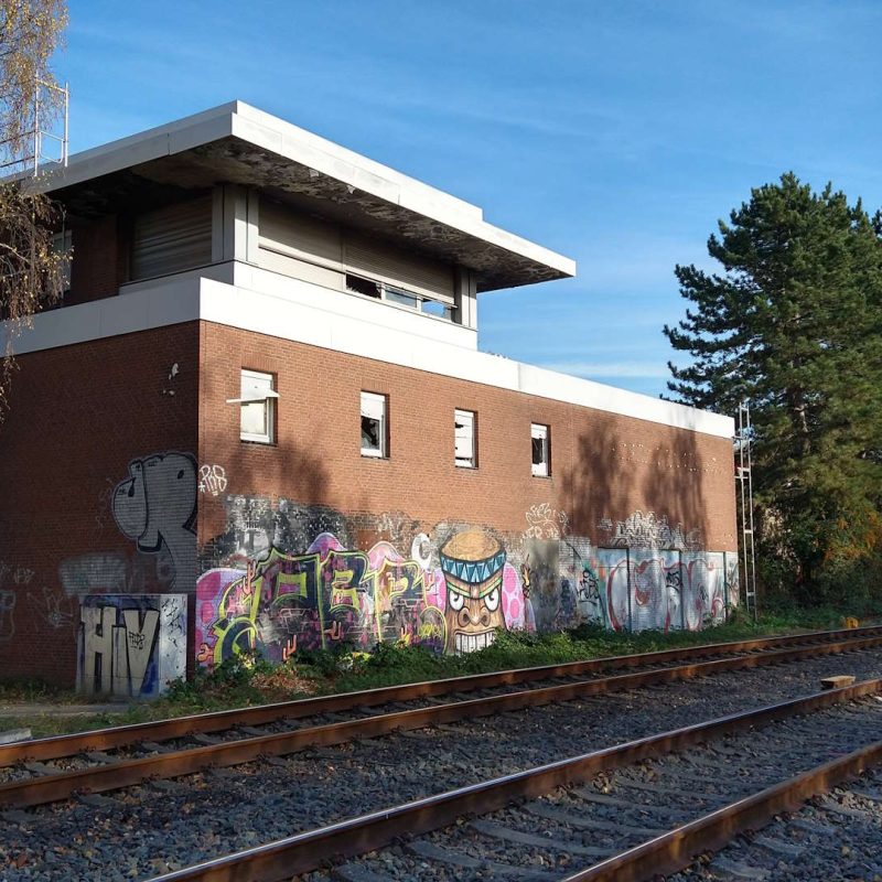 Ein graffitidekoriertes Gebäude neben Schienen, mit einem Baum links und einer Person in gelber Weste, die an der Wand steht