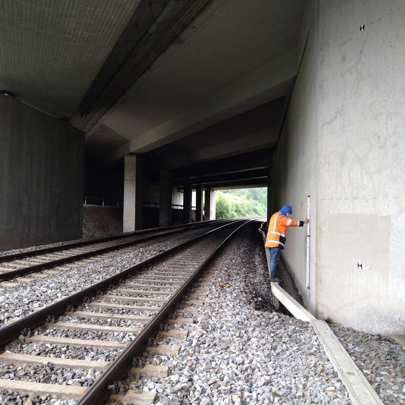 Ein Arbeiter in orangefarbener Sicherheitsjacke misst eine Wand unter einer Brücke neben einem Gleis
