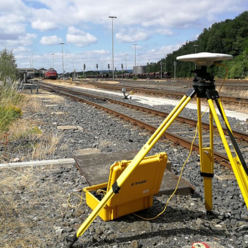 Ein gelbes Vermessungsgerät steht auf einem Gleis, umgeben von Gras und Schotter, mit einem Bahnhof im Hintergrund