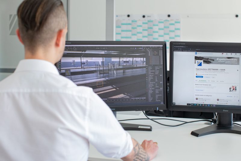 Eine Person in einem weißen Hemd sitzt vor zwei Computermonitoren, einer zeigt eine Grafik eines Bahnhofs, der andere eine Webseite