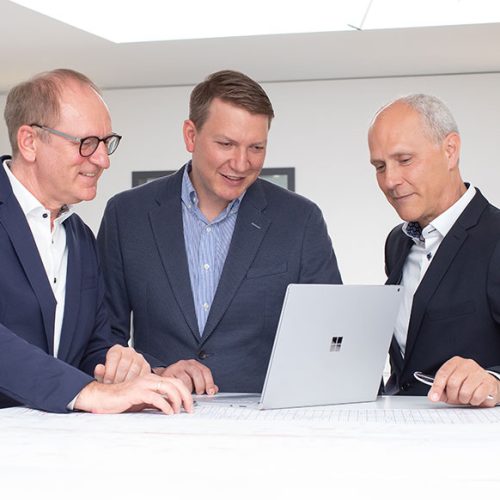 Drei Männer stehen um einen Tisch, schauen auf einen Laptop und diskutieren Pläne, umgeben von modernem Raumdesign